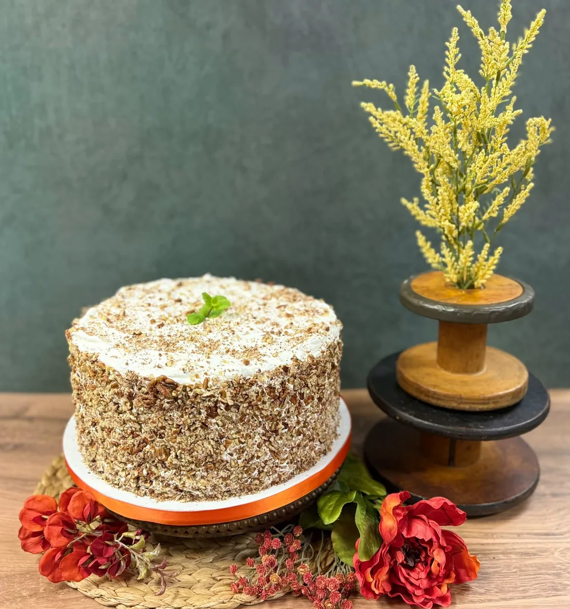 A carrot cake coated in chopped pecans on a cake stand, with red flowers and a spool vase of yellow blooms