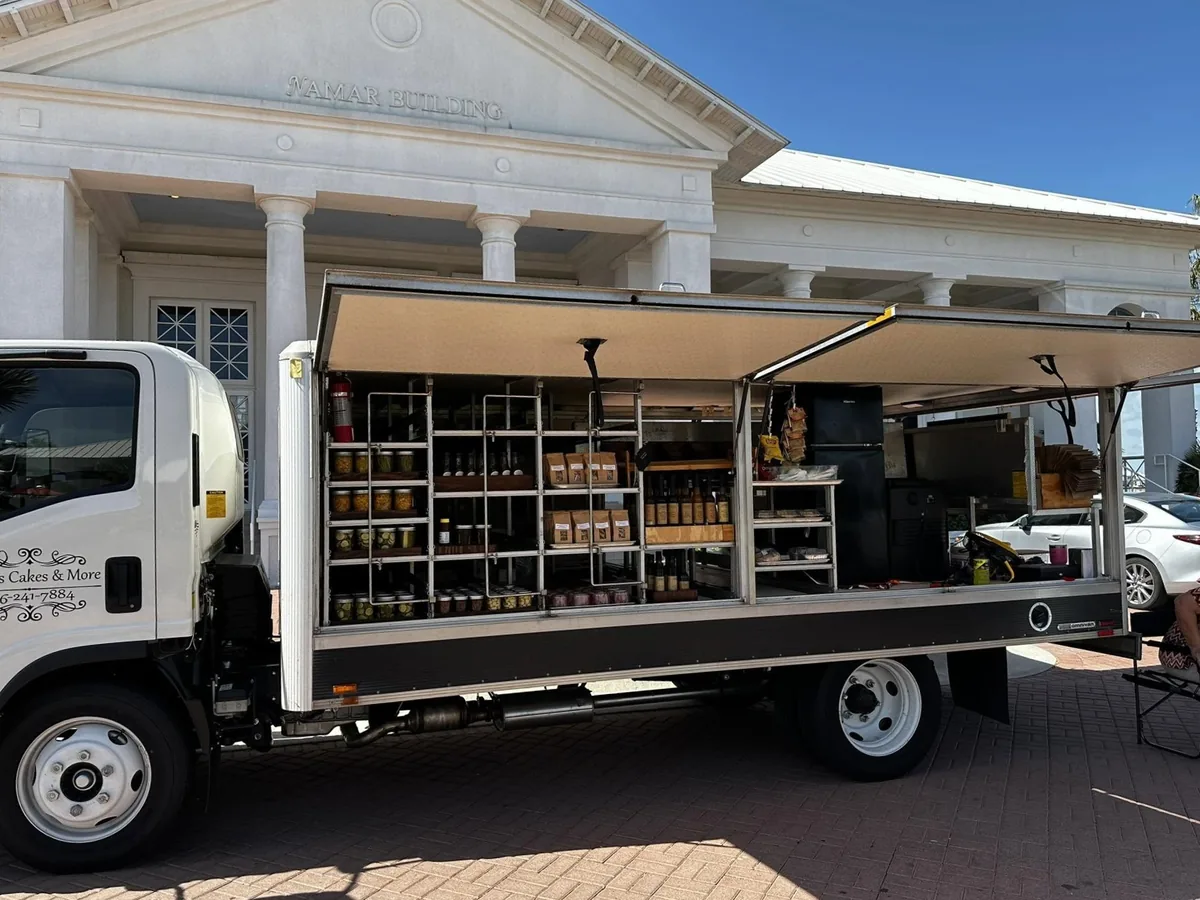 Peggy's Cakes & More truck with its side open, showing shelves of jars, sauces and baked goods