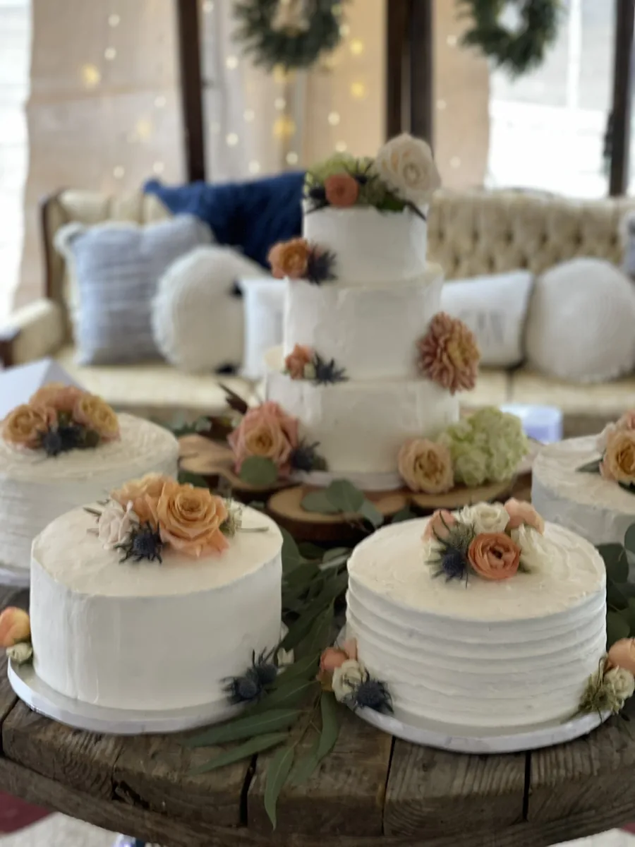 A three-tier white wedding cake with fresh flowers, surrounded by matching single-tier cakes on wood slices