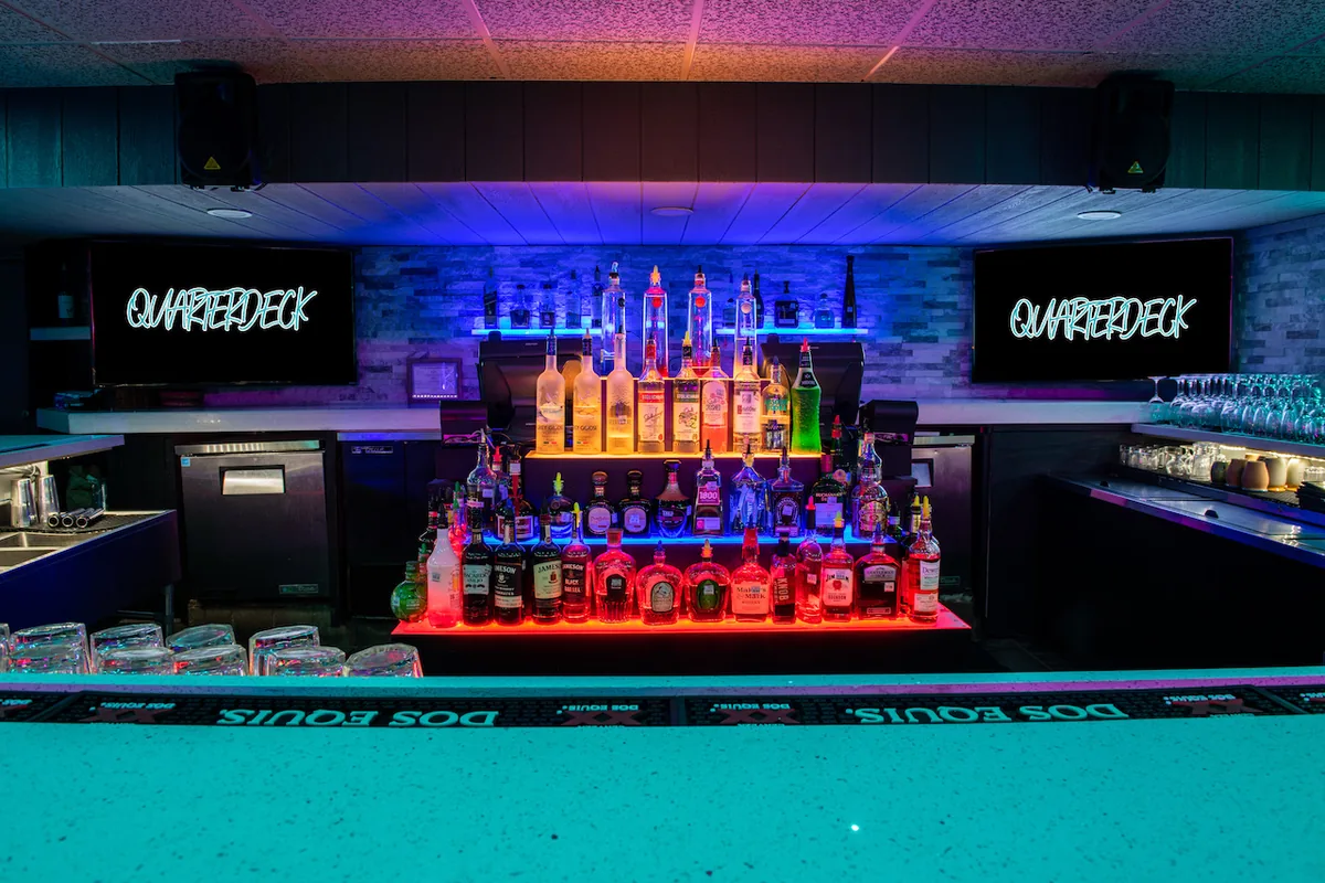 The Quarterdeck bar: backlit shelves of bottles between two QUARTERDECK neon signs, above a turquoise bar top