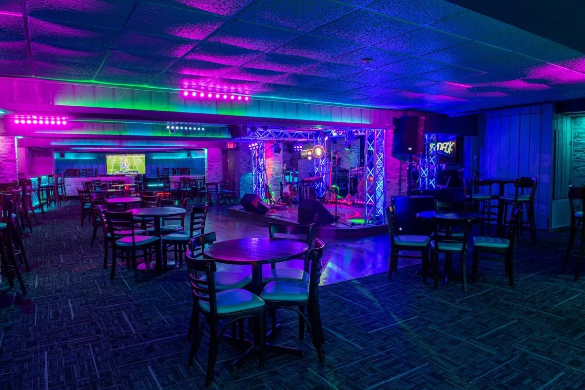 The lounge lit in purple and blue, with high-top tables and chairs, TVs and the stage at the back