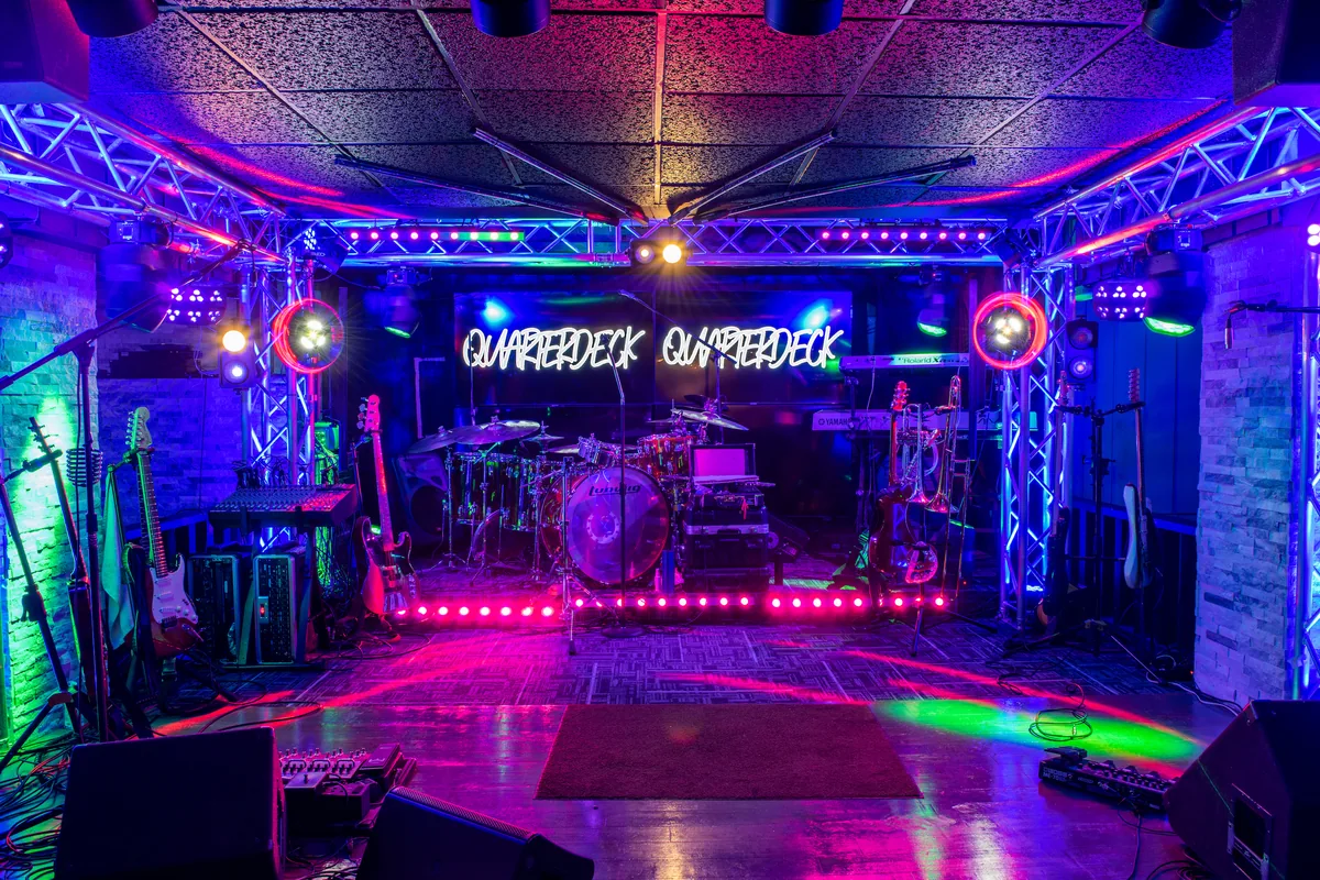 The band stage under a lighting truss with QUARTERDECK neon signs, a drum kit, guitars and the dance floor