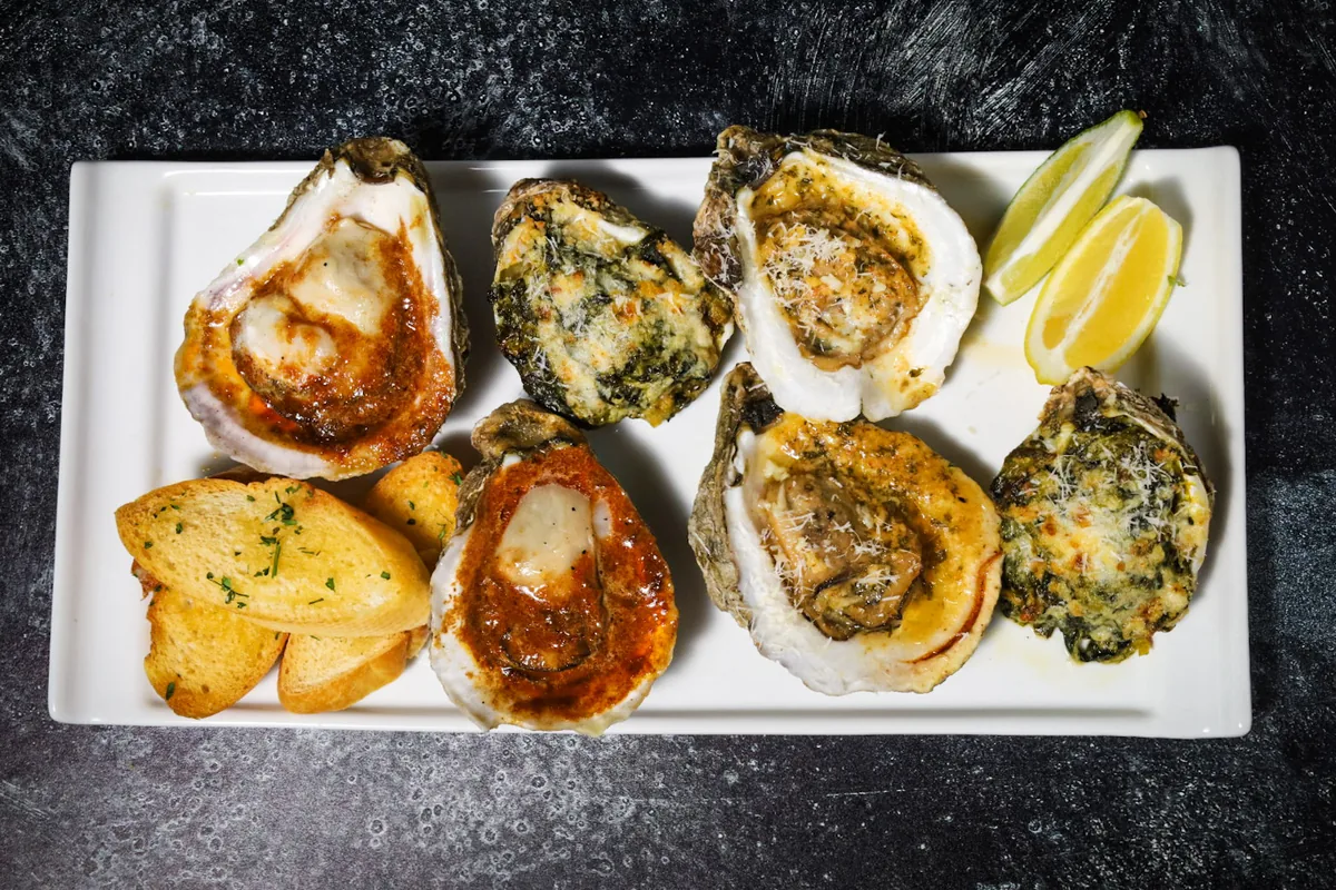 Baked oysters with different toppings (spinach, red sauce, garlic butter) on a long white plate with garlic toast and lemon