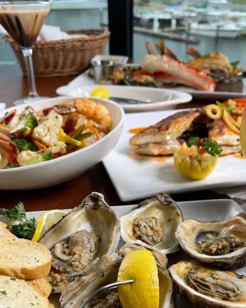 A table by the window set with oysters, a seafood salad, grilled fish and king crab, with the marina outside