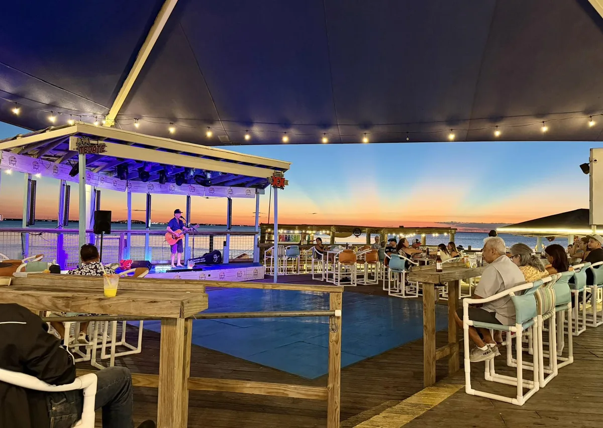 A musician on the stage and guests at tables under the big blue canopy as the sun sets over the bay