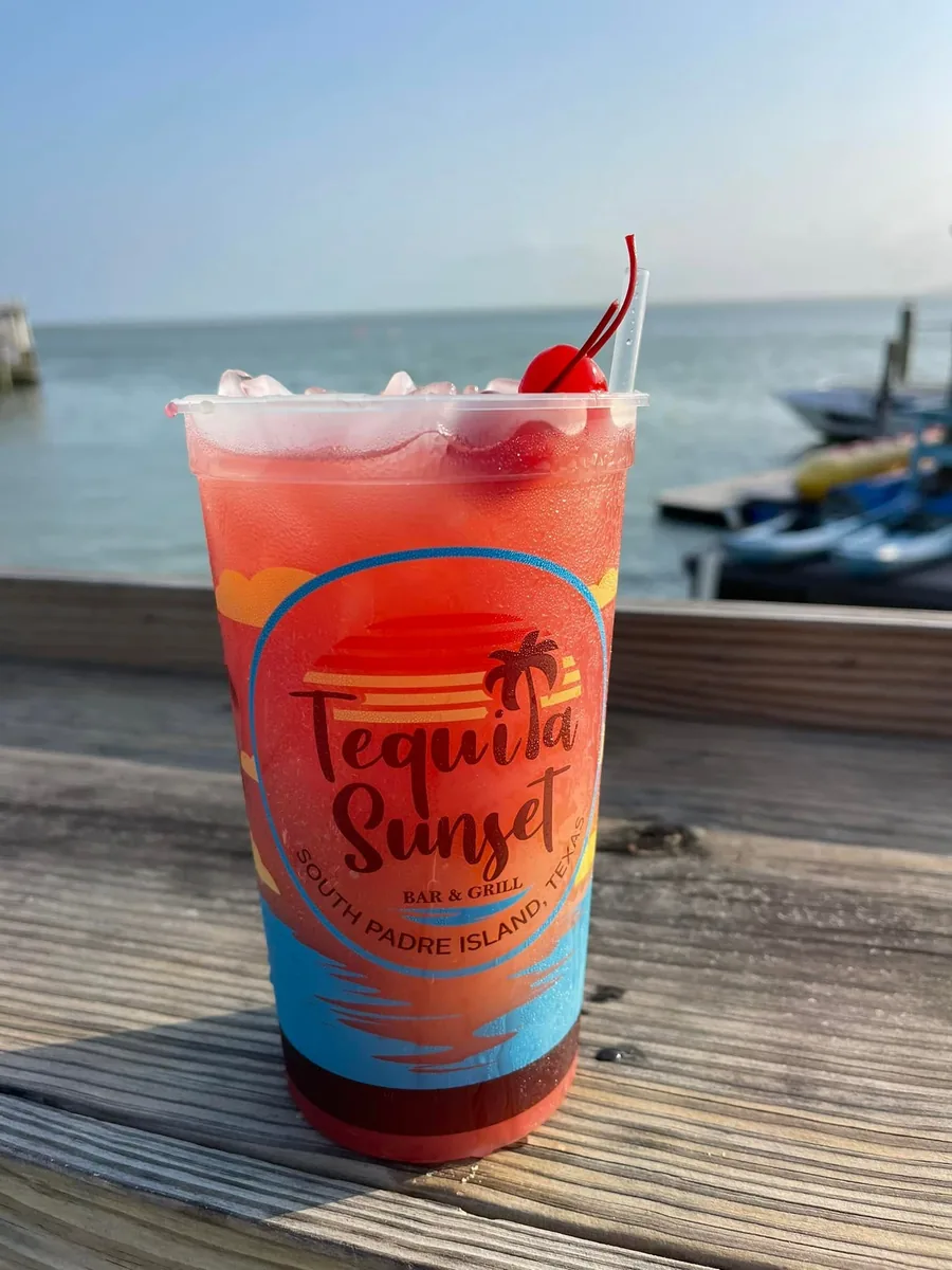 A red frozen drink with a cherry in a Tequila Sunset cup on the rail, with the bay behind
