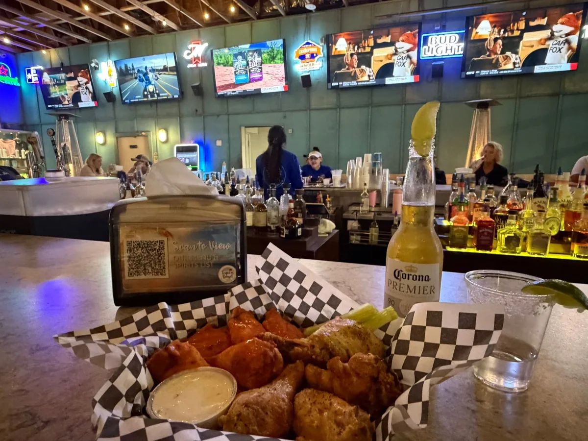 A basket of wings with ranch and celery and a Corona at the bar, under a row of TVs