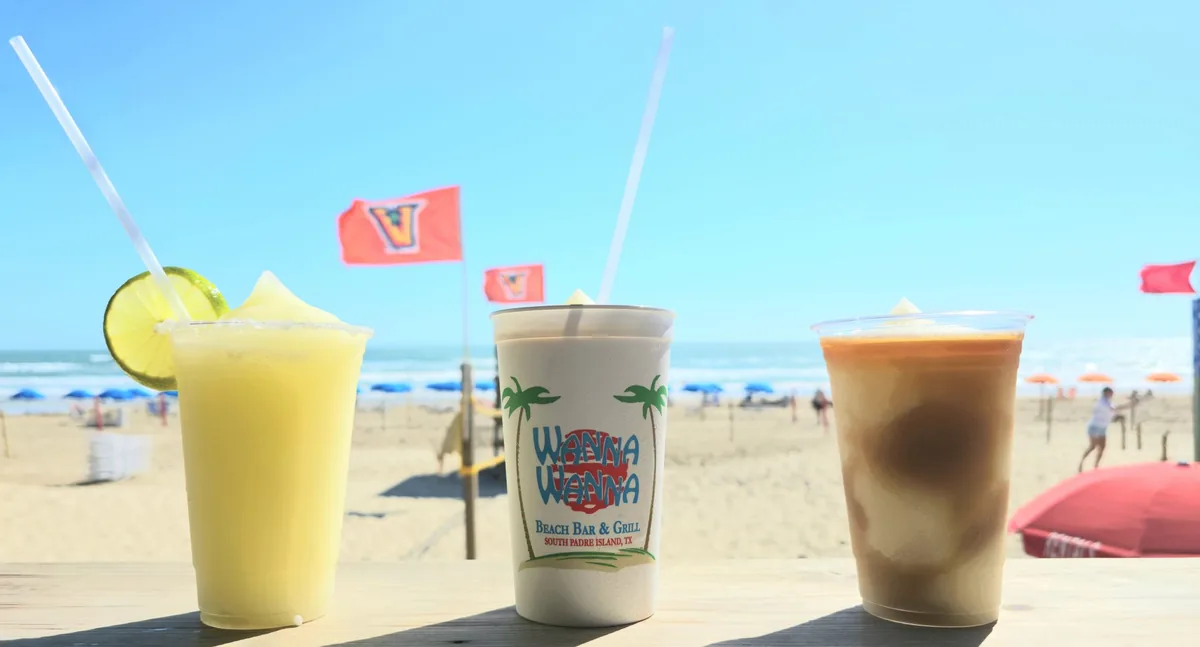 Three frozen drinks on the deck rail with the beach, umbrellas and flags behind