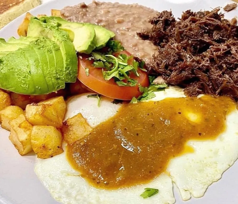 Pulled barbacoa with two fried eggs under green sauce, breakfast potatoes, refried beans, avocado and tomato
