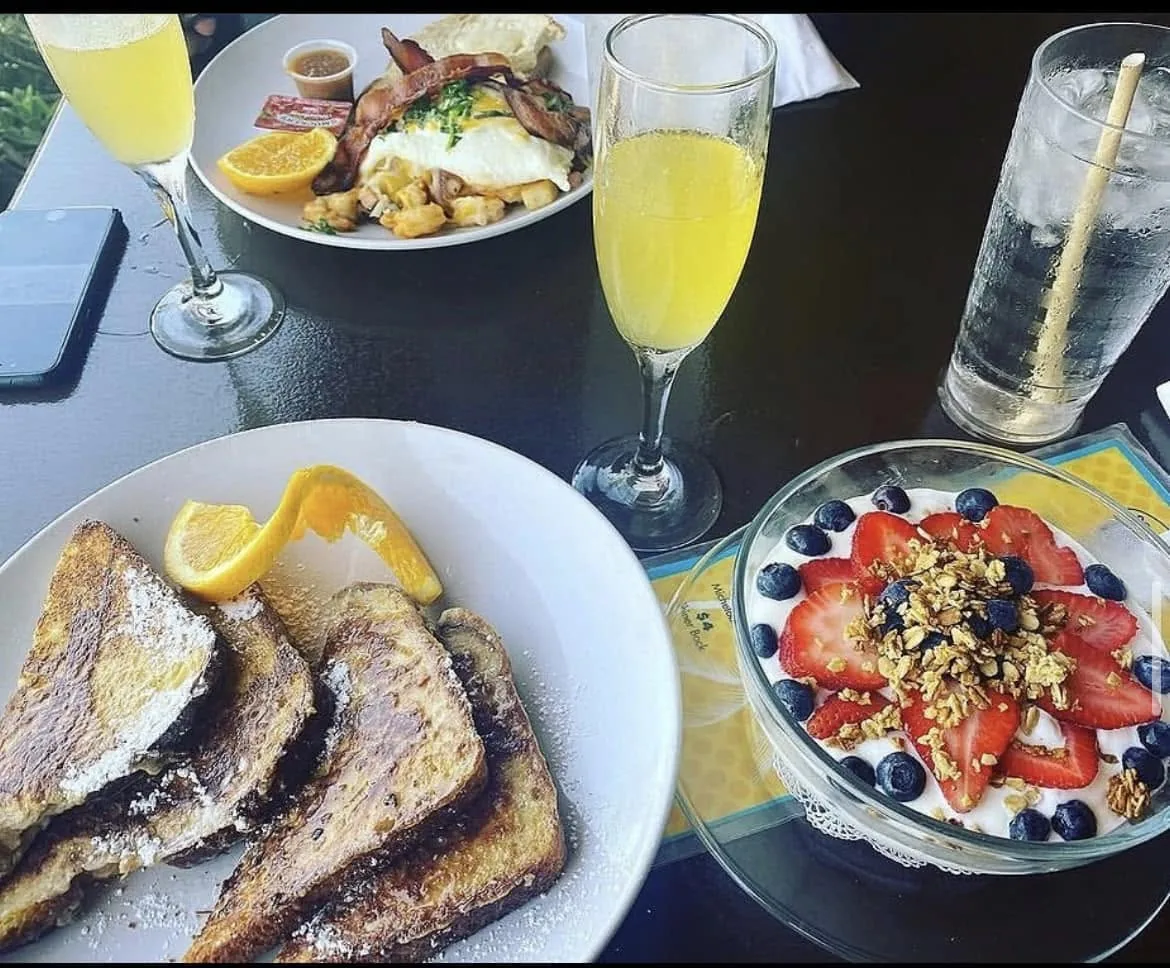 A brunch table on the patio: French toast, a yogurt parfait with berries and granola, a plate of eggs, bacon and potatoes, and mimosas