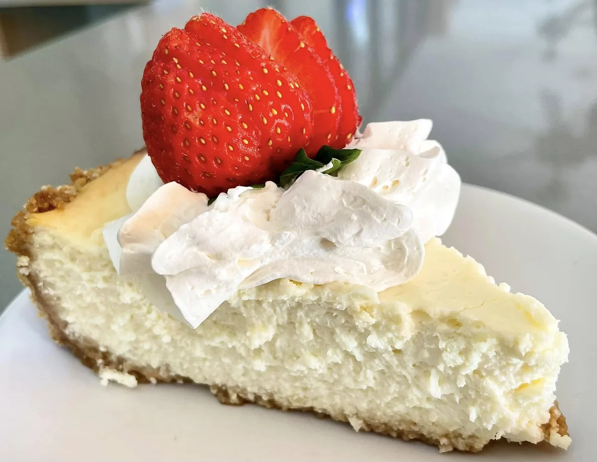 A slice of cheesecake on a graham crust topped with whipped cream and a fanned strawberry
