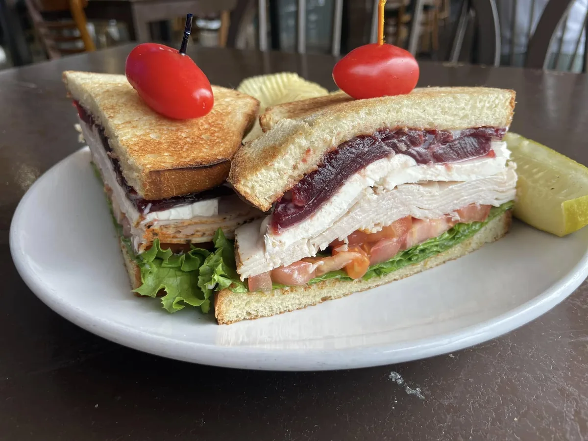 A double-decker sandwich of sliced turkey, cranberry sauce, cream cheese, lettuce and tomato on toast, with chips and a pickle spear